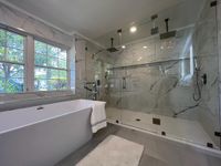 Luxurious Bathroom with Freestanding Tub and Marble Shower: A spacious bathroom featuring a freestanding white bathtub, a glass-enclosed shower with marble tiles, and large windows allowing natural light to enhance the modern design.