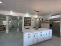 Contemporary kitchen design featuring a large central island with white quartz countertops, elegant pendant lighting, open wooden shelves, and a scenic view of a sparkling outdoor pool through sliding glass doors.